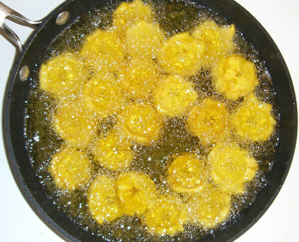 Rodajas de plátano verde (tostones) friéndose en aceite caliente en una sartén negra.