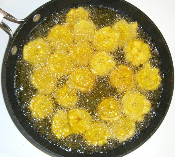 Rodajas de plátano verde (tostones) friéndose en aceite caliente en una sartén negra.