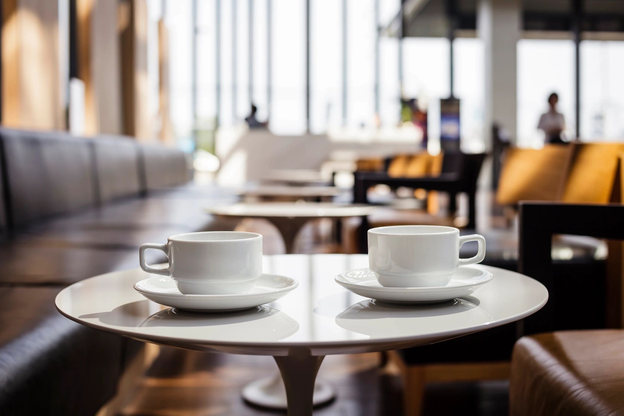 Dos tazas de café blancas sobre platillos, situadas sobre una mesa redonda en una cafetería acogedora con decoración moderna y asientos de cuero.