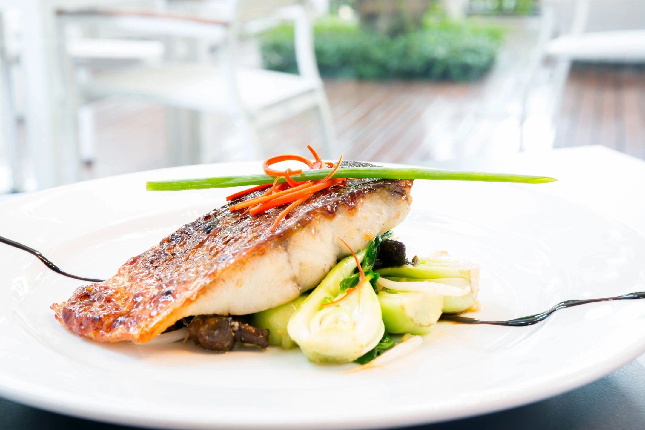 Filete de pescado a la parrilla servido en un plato blanco, acompañado de vegetales salteados y decorado con tiras de zanahoria y una hoja de cebollín.