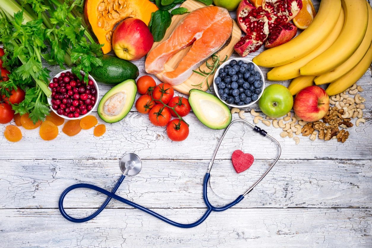 Una variedad de frutas frescas y salmón dispuestos alrededor de un estetoscopio con un corazón rojo, sobre un fondo de madera blanca.