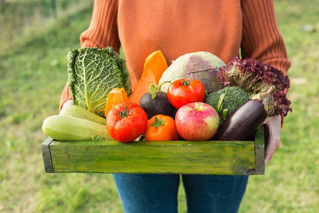 Una persona sostiene una caja de madera llena de vegetales frescos y coloridos en un entorno al aire libre.