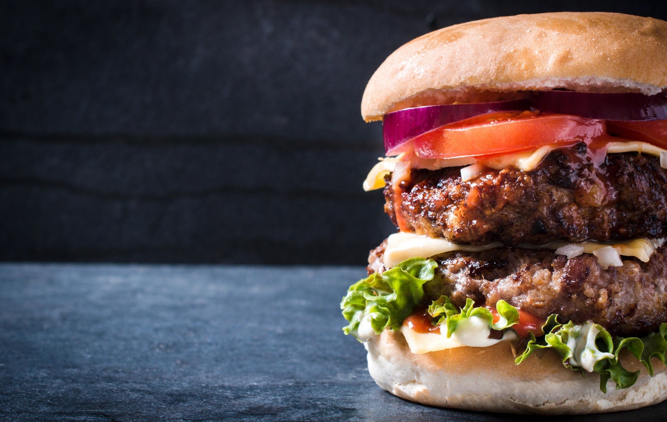 Una hamburguesa doble con queso, lechuga, tomate y cebolla sobre un fondo oscuro.