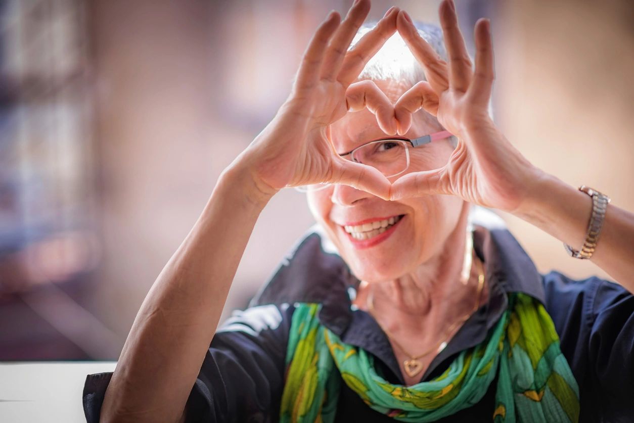 Una mujer mayor sonriente haciendo un corazón con las manos alrededor de su rostro.