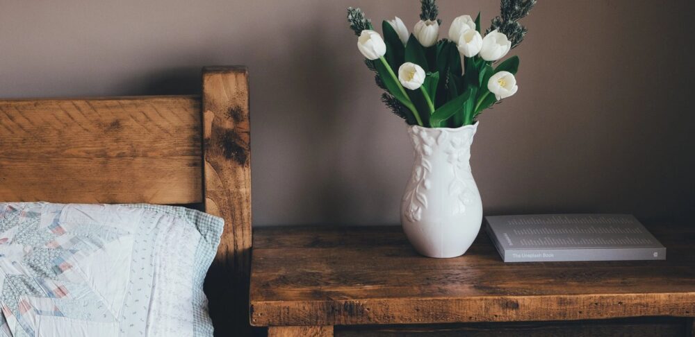 Un jarrón con flores blancas en una mesa de madera al lado de una cama.