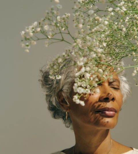 Una mujer mayor con el pelo canoso cerrando los ojos y disfrutando del aroma de unas flores blancas delicadas.