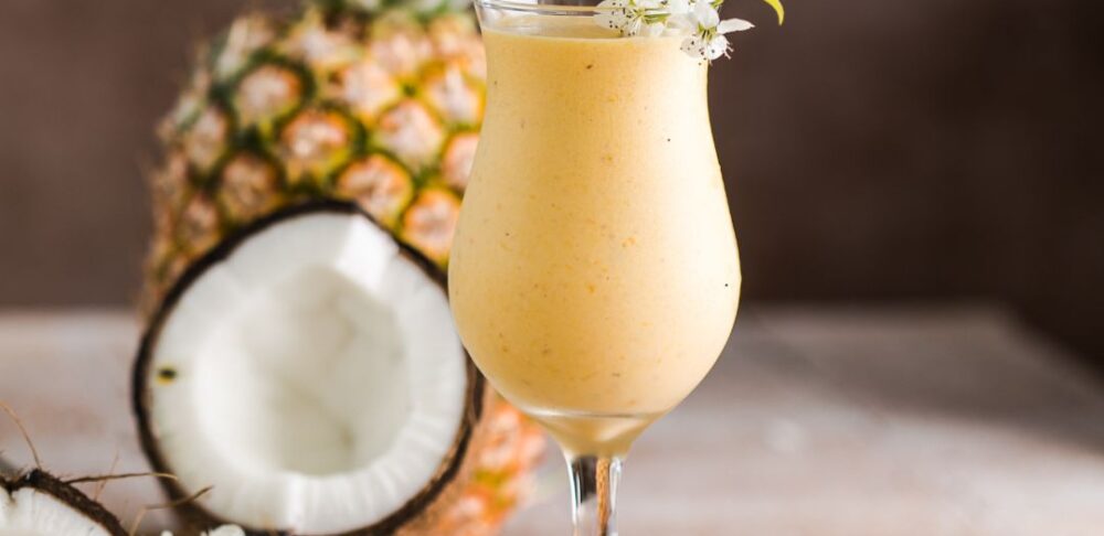 Batido tropical de piña y coco en una copa, adornado con flores y al fondo una piña y coco frescos.