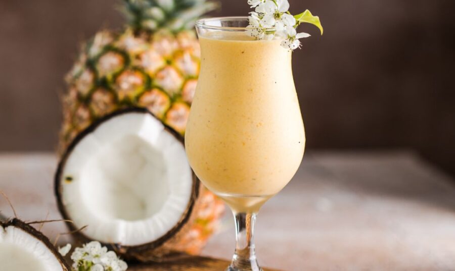 Batido tropical de piña y coco en una copa, adornado con flores y al fondo una piña y coco frescos.