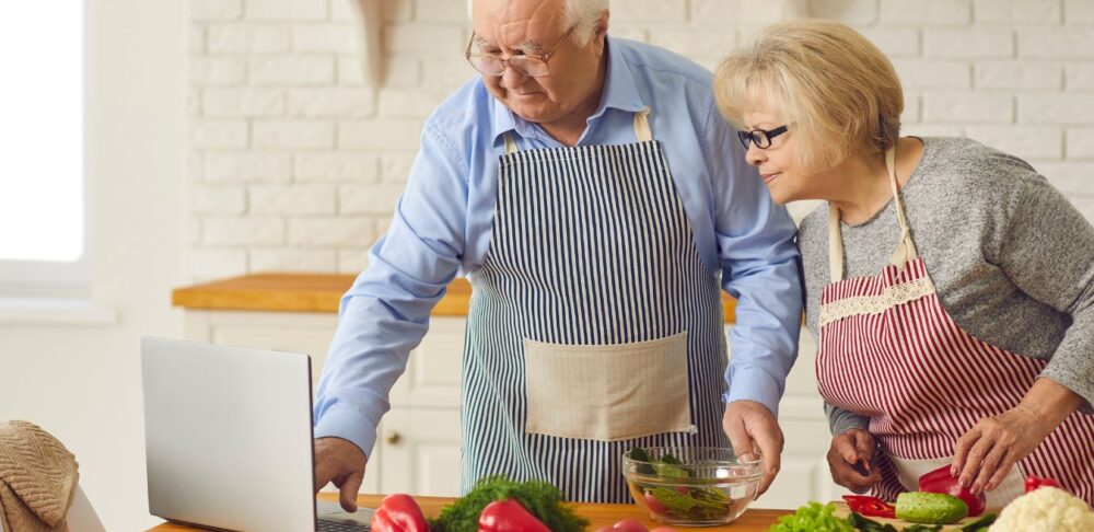 Pareja de adultos mayores buscando recetas en línea mientras cocinan juntos.