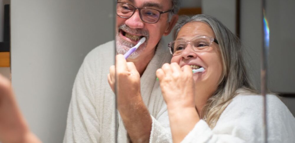 Una pareja mayor sonriente, usando batas de baño blancas, cepillándose los dientes juntos frente a un espejo.