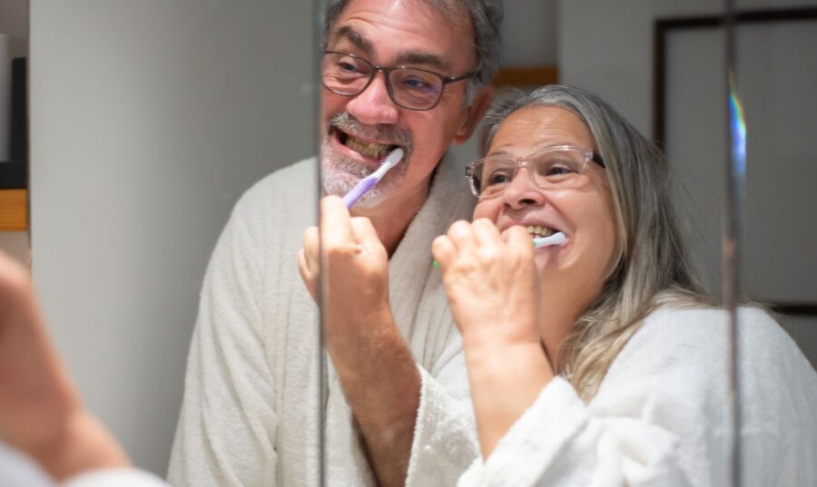 Una pareja mayor sonriente, usando batas de baño blancas, cepillándose los dientes juntos frente a un espejo.