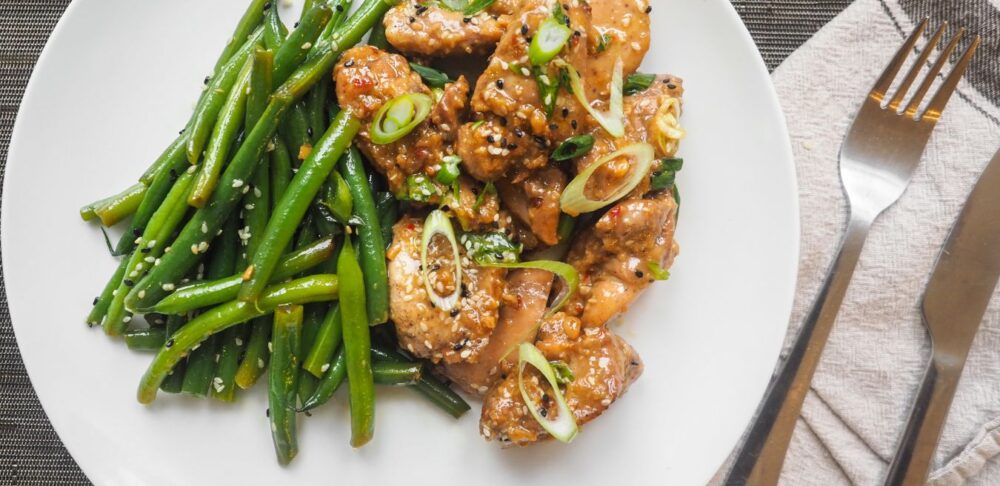 Plato de pollo con semillas de sésamo y judías verdes en un plato blanco, presentado en una mesa con mantel y cubiertos.