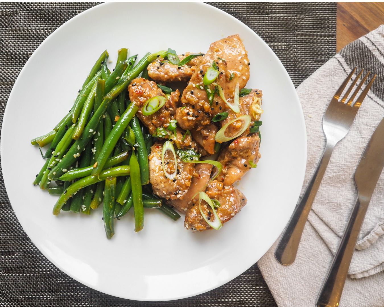 Plato de pollo con semillas de sésamo y judías verdes en un plato blanco, presentado en una mesa con mantel y cubiertos.