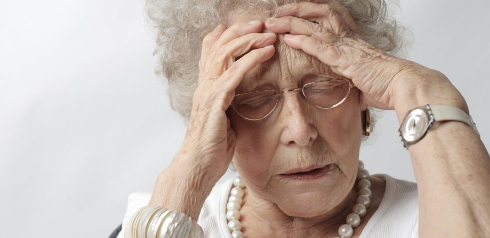 Una mujer mayor con joyería elegante, expresando preocupación o dolor de cabeza, con ambas manos en la frente y los ojos cerrados.