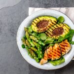 Plato saludable con aguacate a la parrilla, espárragos verdes, pechuga de pollo a la parrilla y semillas de lino, servido en una mesa de piedra gris con una servilleta de lino.