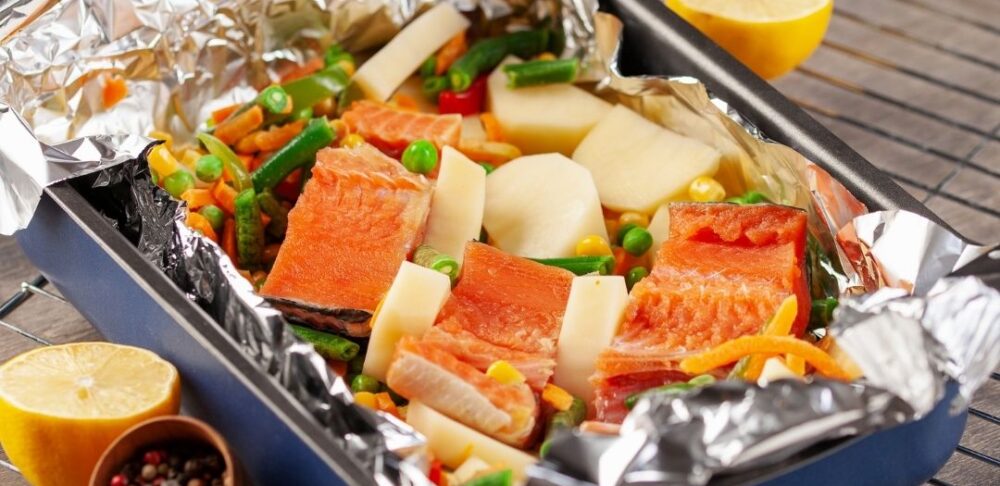 Una bandeja para horno con salmón y vegetales mixtos envuelta en papel de aluminio. El plato incluye judías verdes, guisantes y trozos de patatas, y se coloca sobre una rejilla para enfriar. También hay mitades de limón y un tazón pequeño de granos de pimienta mezclados al lado del plato.