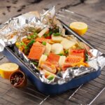 Una bandeja para horno con salmón y vegetales mixtos envuelta en papel de aluminio. El plato incluye judías verdes, guisantes y trozos de patatas, y se coloca sobre una rejilla para enfriar. También hay mitades de limón y un tazón pequeño de granos de pimienta mezclados al lado del plato.