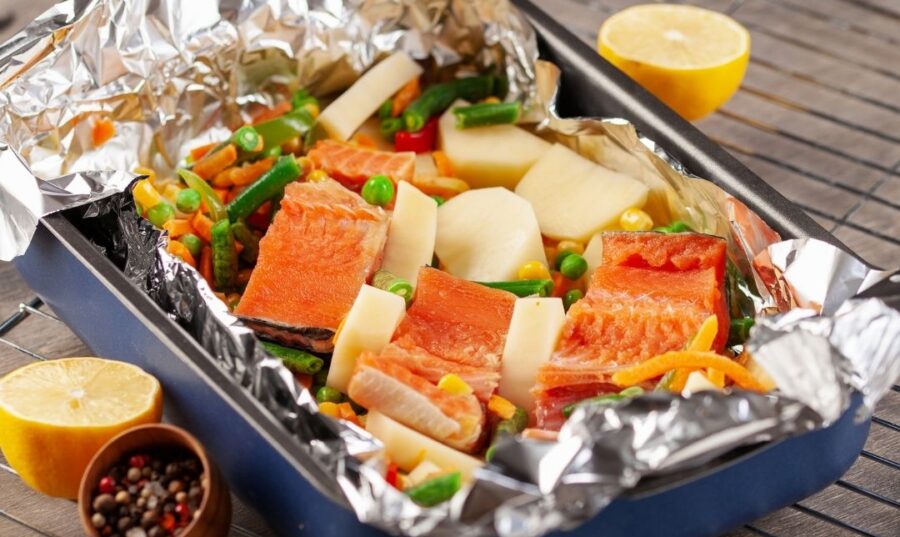 Una bandeja para horno con salmón y vegetales mixtos envuelta en papel de aluminio. El plato incluye judías verdes, guisantes y trozos de patatas, y se coloca sobre una rejilla para enfriar. También hay mitades de limón y un tazón pequeño de granos de pimienta mezclados al lado del plato.