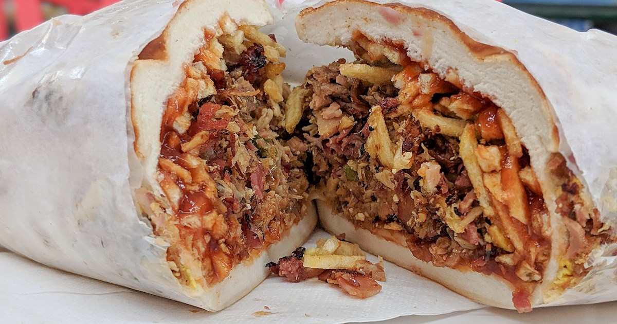 Un sándwich Tripleta rebanado por la mitad, mostrando un relleno abundante de carne, papas fritas y salsa, envuelto en papel blanco.