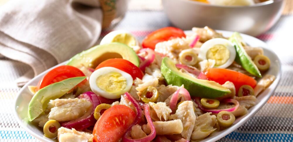 Ensalada de bacalao con aguacate, tomate, huevo cocido, cebolla roja y aceitunas verdes en un plato blanco, sobre una mesa de madera con un mantel colorido.