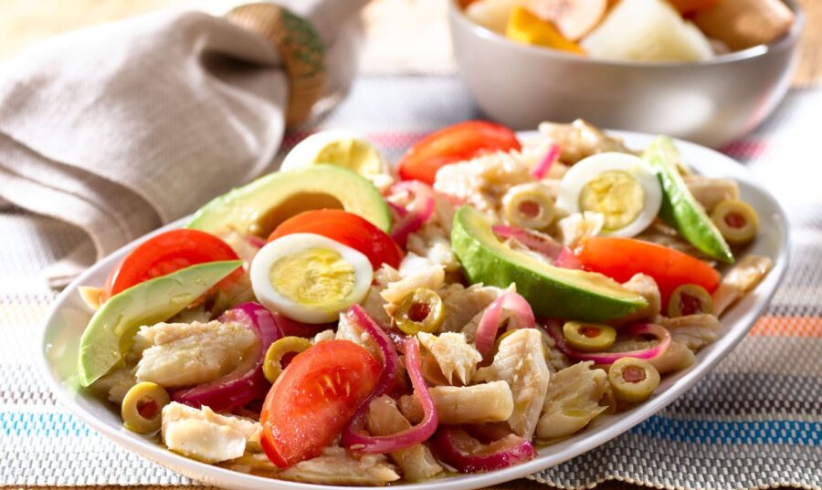 Ensalada de bacalao con aguacate, tomate, huevo cocido, cebolla roja y aceitunas verdes en un plato blanco, sobre una mesa de madera con un mantel colorido.