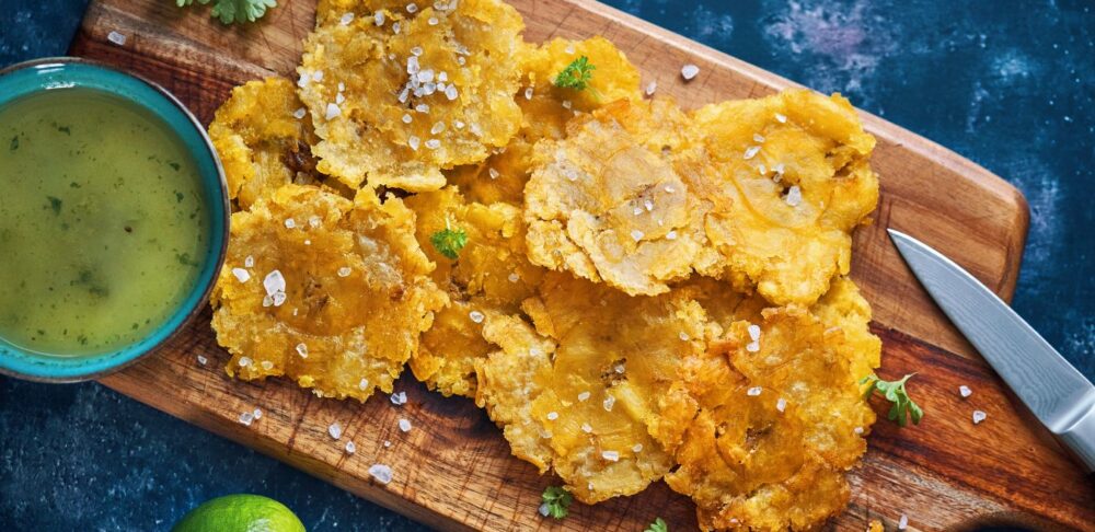 Plato de Tostones servidos en una tabla de cortar de madera, acompañados de una salsa verde en un tazón y una rodaja de limón.