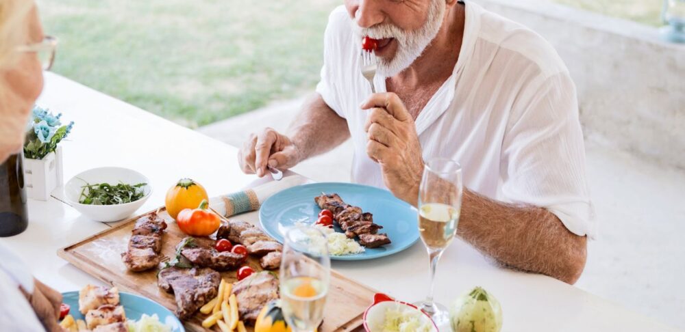 Pareja de adultos mayores disfrutando de una comida saludable al aire libre, con platos de carne, ensalada y copas de champaña.