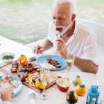 Pareja de adultos mayores disfrutando de una comida saludable al aire libre, con platos de carne, ensalada y copas de champaña.