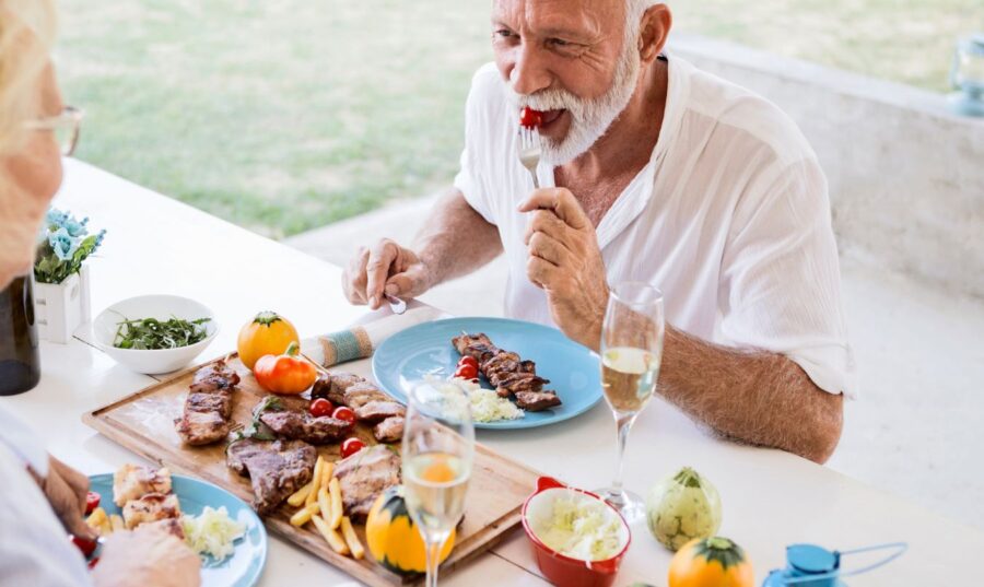 Pareja de adultos mayores disfrutando de una comida saludable al aire libre, con platos de carne, ensalada y copas de champaña.