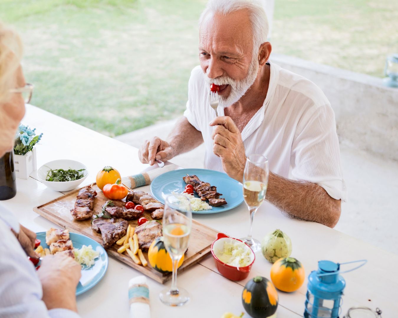 Pareja de adultos mayores disfrutando de una comida saludable al aire libre, con platos de carne, ensalada y copas de champaña.