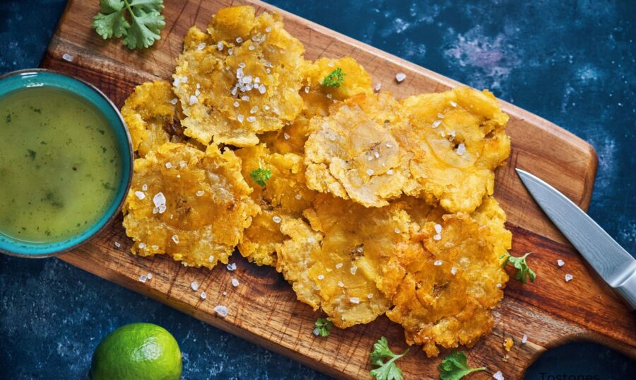 Plato de Tostones servidos en una tabla de cortar de madera, acompañados de una salsa verde en un tazón y una rodaja de limón.