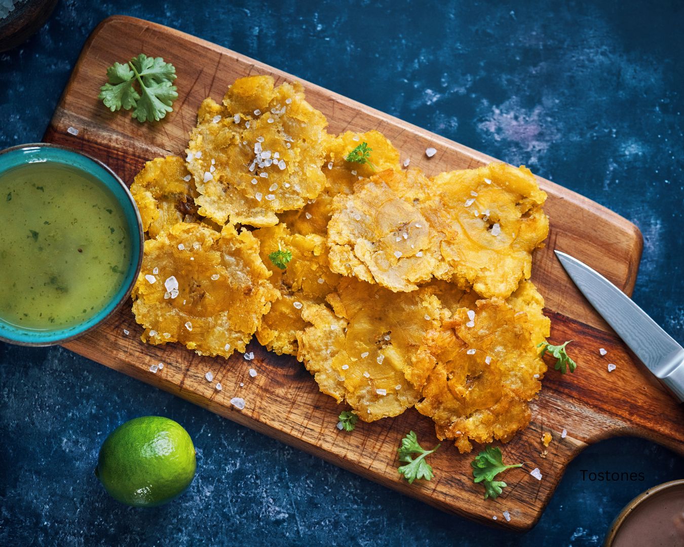 Plato de Tostones servidos en una tabla de cortar de madera, acompañados de una salsa verde en un tazón y una rodaja de limón.