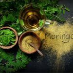 Hojas frescas y polvo de moringa en tazones de madera, junto a una taza de té de moringa, con la palabra "Moringa" escrita en el polvo sobre una superficie oscura.