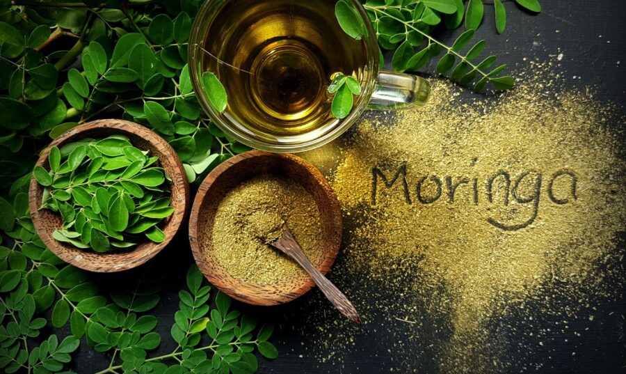 Hojas frescas y polvo de moringa en tazones de madera, junto a una taza de té de moringa, con la palabra "Moringa" escrita en el polvo sobre una superficie oscura.