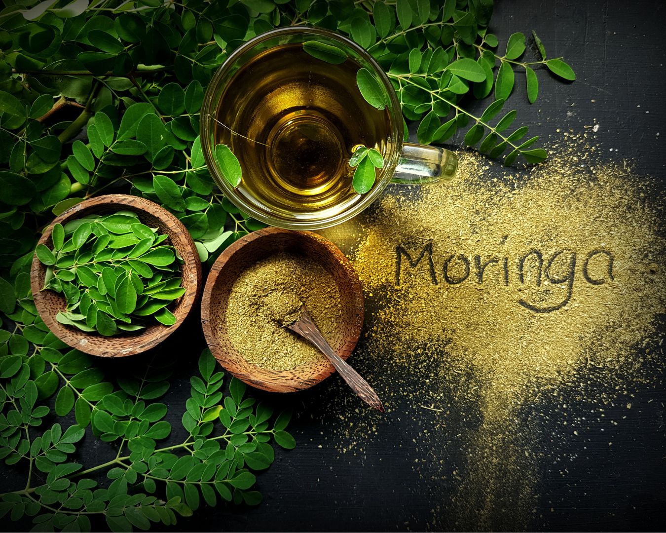 Hojas frescas y polvo de moringa en tazones de madera, junto a una taza de té de moringa, con la palabra "Moringa" escrita en el polvo sobre una superficie oscura.