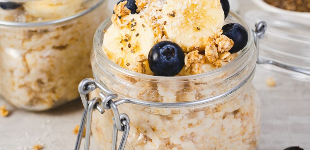 Un tarro de vidrio con avena cocida, insectos comestibles, guineo en rodajas, arándanos y granola, sobre una mesa de madera clara.