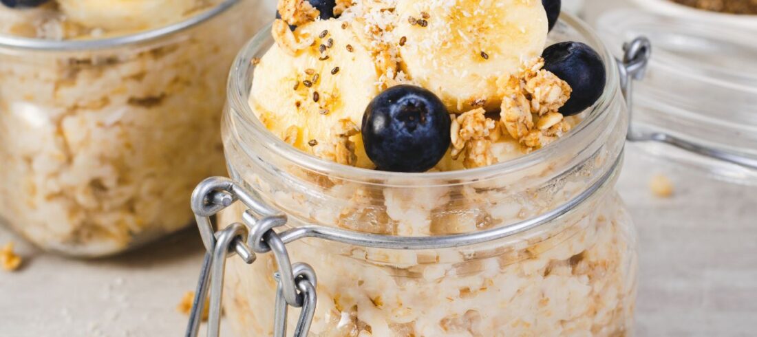 Un tarro de vidrio con avena cocida, insectos comestibles, guineo en rodajas, arándanos y granola, sobre una mesa de madera clara.