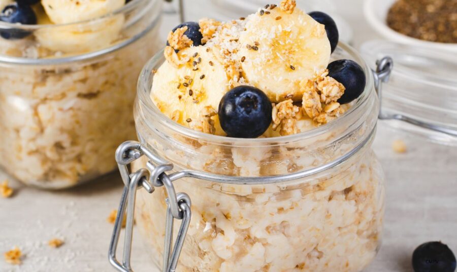 Un tarro de vidrio con avena cocida, insectos comestibles, guineo en rodajas, arándanos y granola, sobre una mesa de madera clara.