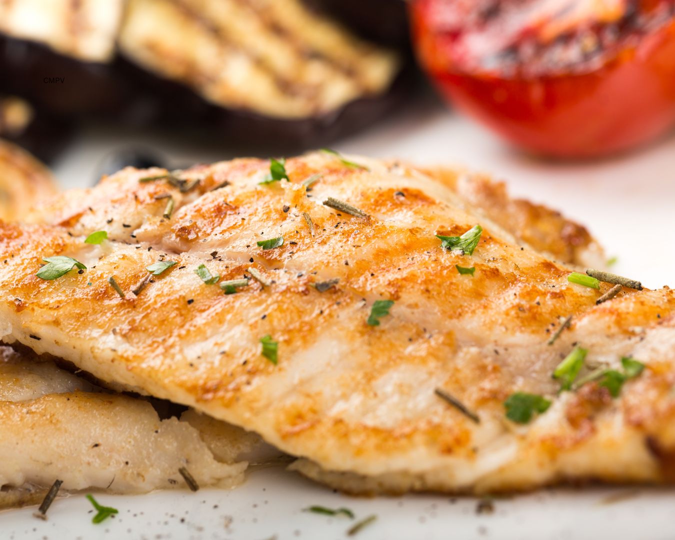 Primer plano de un filete de pescado a la parrilla, adornado con hierbas frescas y acompañado de vegetales a la parrilla en el fondo.