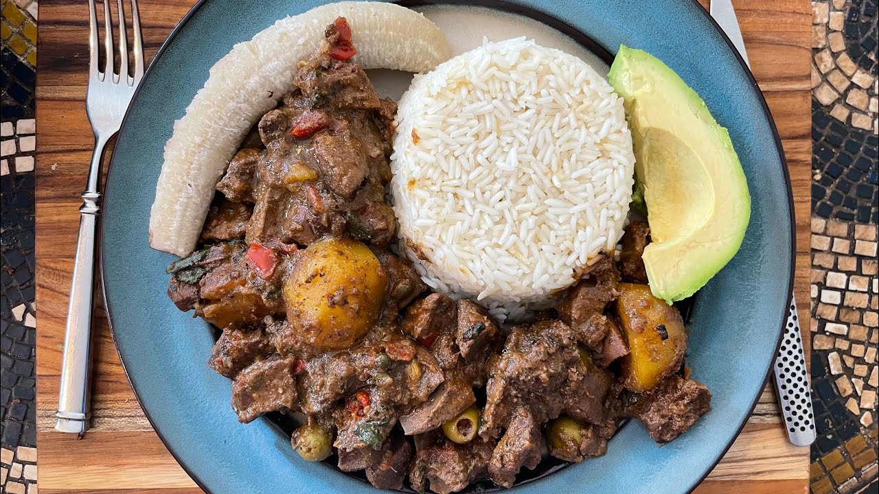 Plato color azul con gandinga guisada con papas y aceitunas. Acompañada con arroz blanco, guineo verde hervido y una rebanada de aguacate.