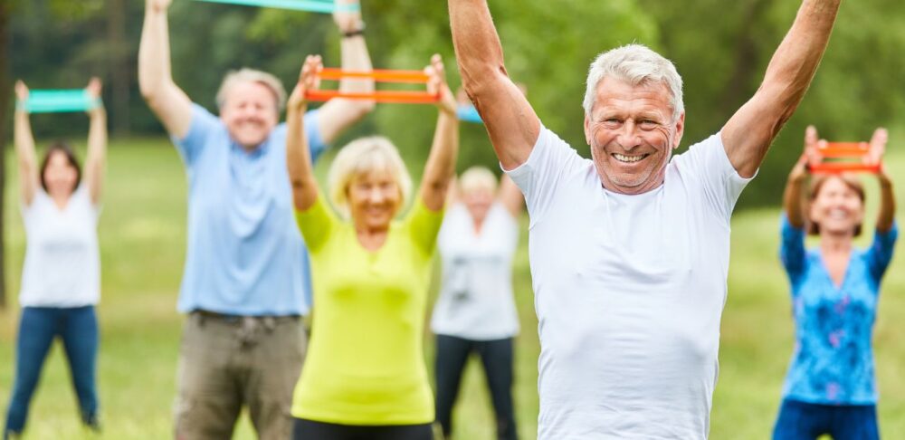 Grupo de adultos mayores realizando ejercicio al aire libre usando bandas de resistencia, mejorando su fuerza y flexibilidad.