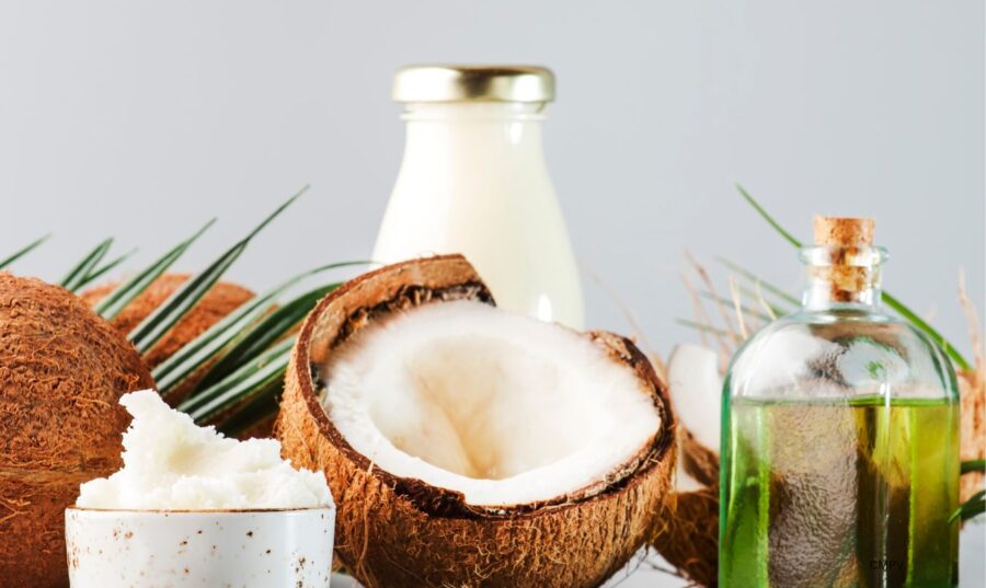 Un coco partido junto a aceite de coco en una botella de vidrio, un tarro con crema de coco y un frasco de leche sobre un fondo claro.