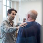 Un médico con camisa gris conversa sobre los eructos excesivos con un adulto mayor, tocándole el hombro en un gesto de apoyo, en un entorno moderno y luminoso.