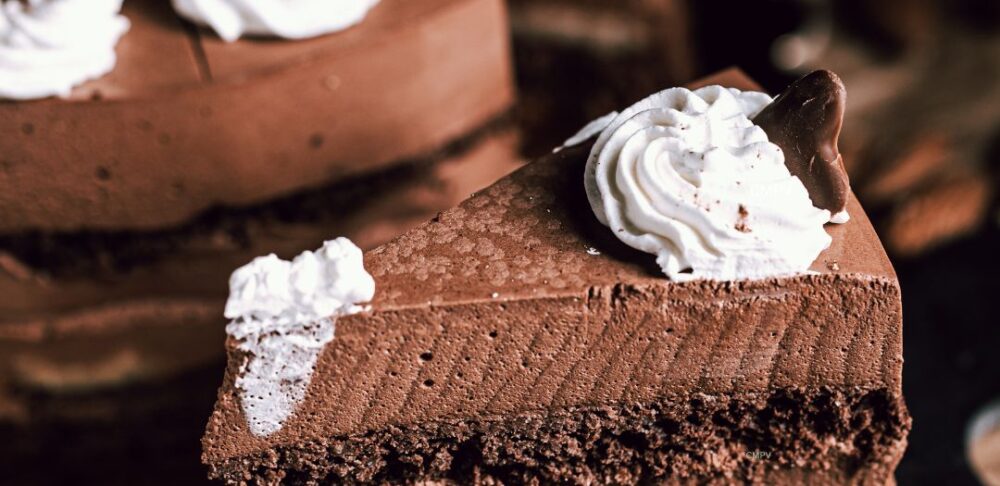 Un apetitoso pastel de chocolate con capas cremosas y decorado con crema batida, colocado en un plato oscuro junto a un tenedor y almendras, capturando un antojo irresistible de algo dulce.