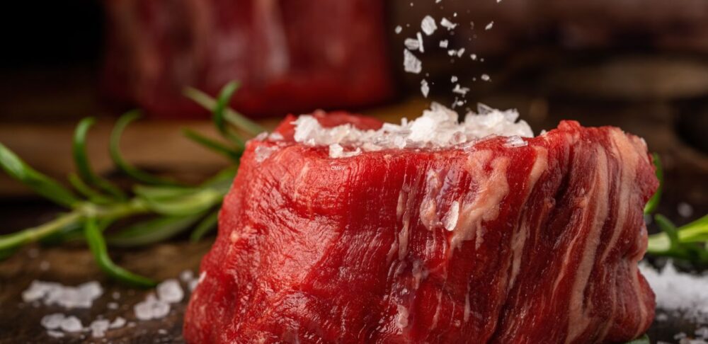 Trozo de carne roja cruda con sal gruesa esparcida encima, sobre fondo rústico con ramas de romero.