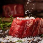 Trozo de carne roja cruda con sal gruesa esparcida encima, sobre fondo rústico con ramas de romero.