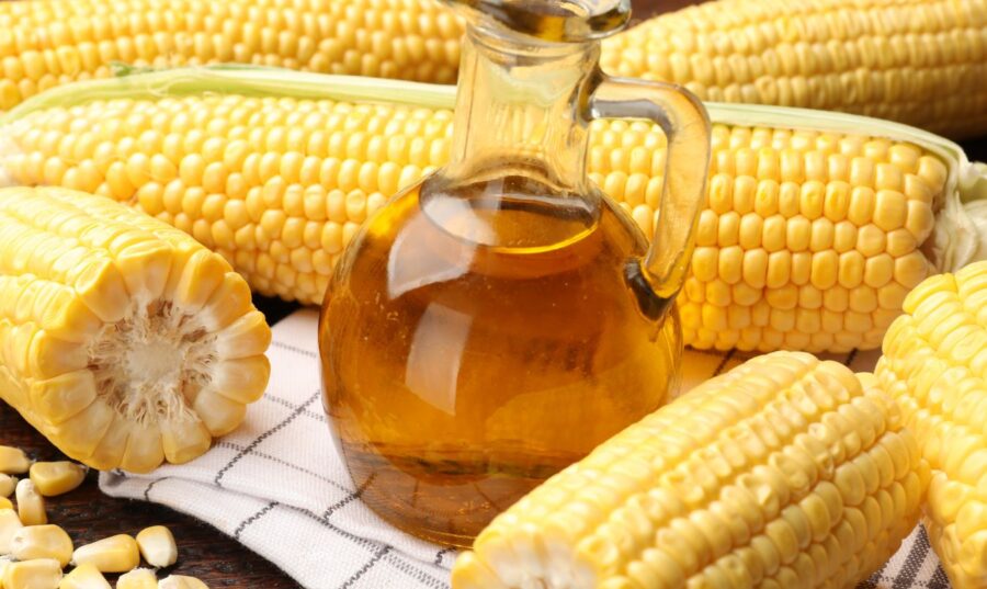 Jarabe de maíz alto en fructosa en frasco, rodeado de mazorcas de maíz, que simboliza el origen del HFCS utilizado en alimentos procesados.