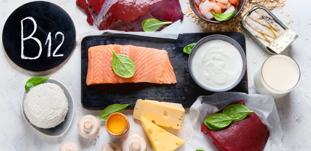 Fuentes alimenticias de vitamina B12 sobre una mesa, incluyendo salmón, hígado, carne de res, camarones, sardinas, leche, yogur, queso, huevo y cereales fortificados.