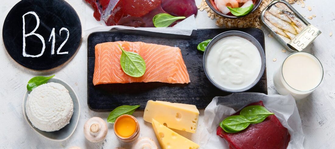 Fuentes alimenticias de vitamina B12 sobre una mesa, incluyendo salmón, hígado, carne de res, camarones, sardinas, leche, yogur, queso, huevo y cereales fortificados.