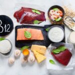 Fuentes alimenticias de vitamina B12 sobre una mesa, incluyendo salmón, hígado, carne de res, camarones, sardinas, leche, yogur, queso, huevo y cereales fortificados.